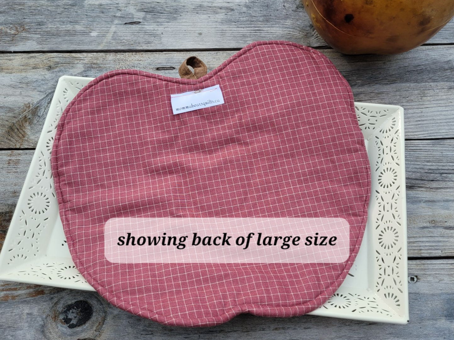 A quilted apple-shaped hot mat in pink and white grid pattern fabric is displayed on a white decorative tray over a rustic gray wooden surface. There is a transluscent label reading 'showing back of large size'.