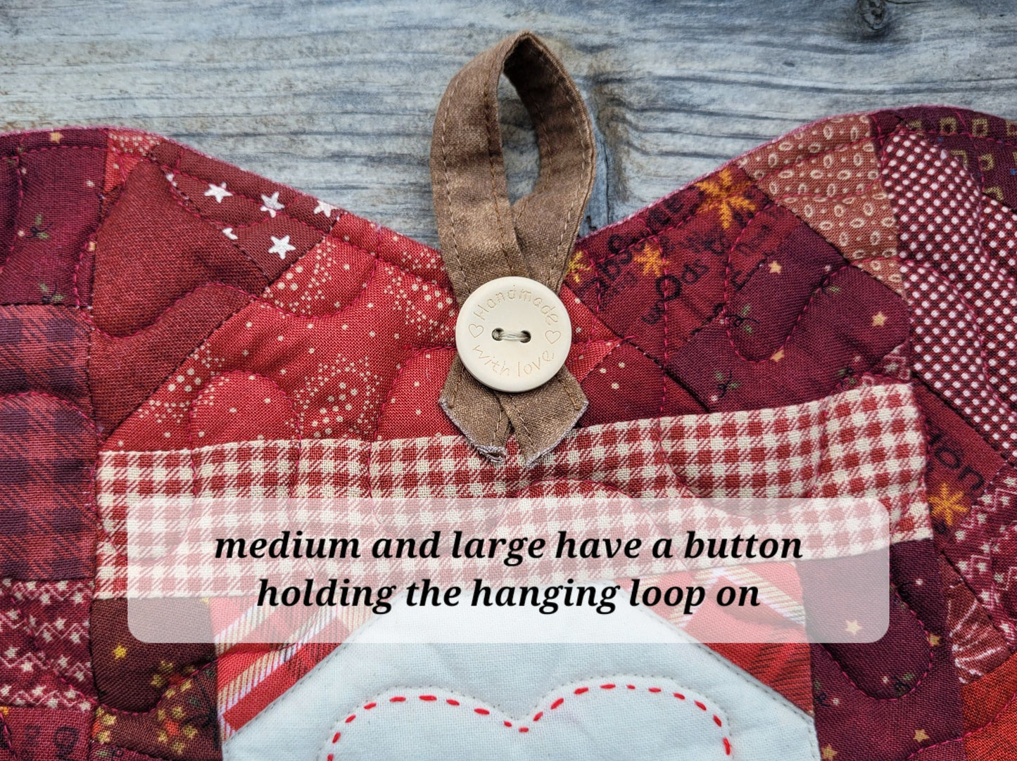 Close-up of a quilted patchwork fabric apple with red and burgundy patterns, a brown hanging loop, and a white button. A translucent text overlay reads, 'medium and large have a button holding the hanging loop on.