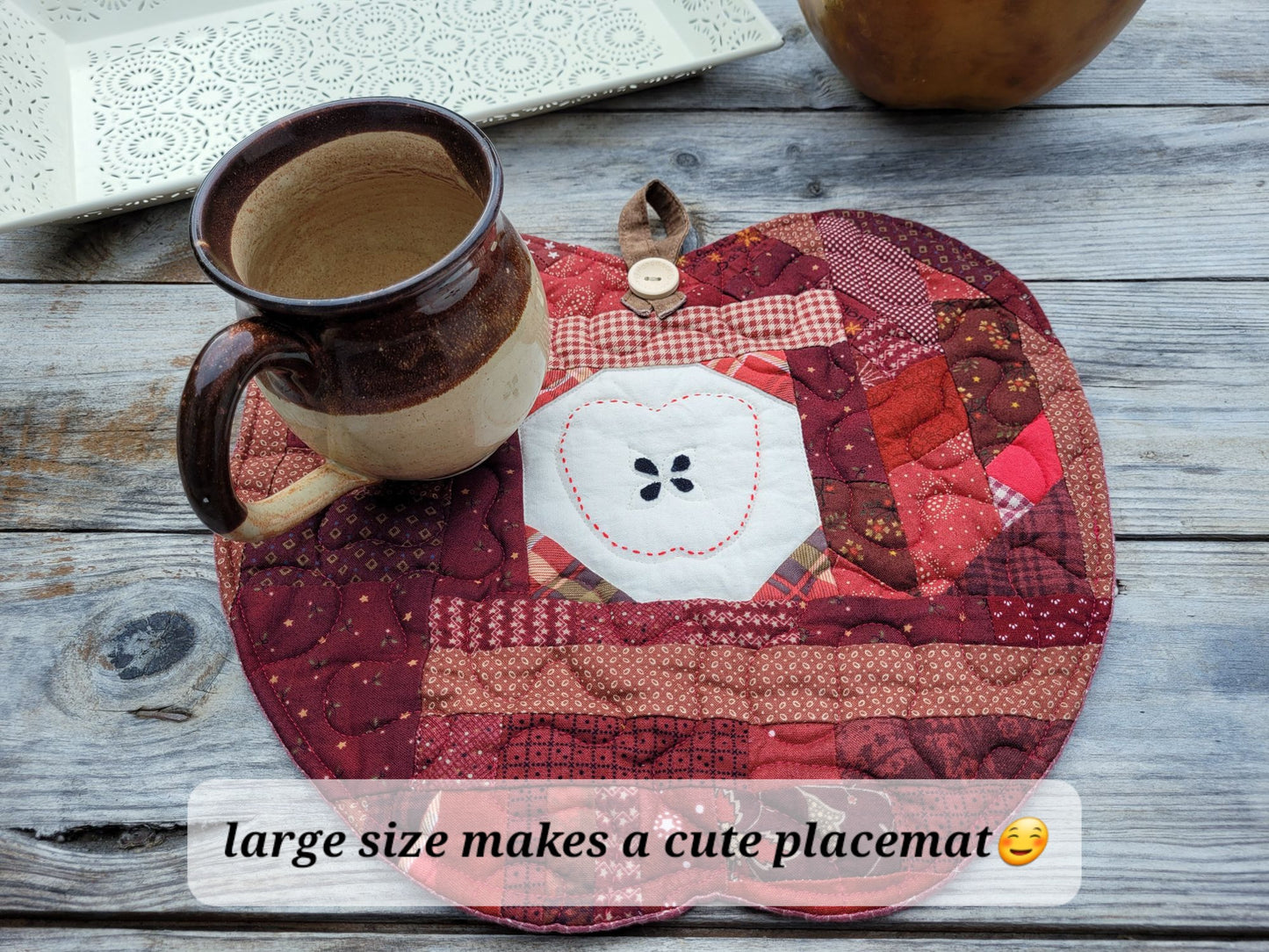 A handmade quilted placemat shaped like an apple, featuring a central white muslin section with hand stitched apple outline around four appliqued seeds, a wooden button at the top of the mat holds the brown hanging loop. The placemat is styled on a rustic gray wooden table with a pottery mug resting on top.