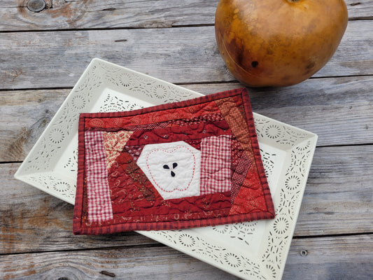 A small red and white patchwork quilted coaster featuring a central white octagonal patch with a handstitched apple motif around three appliqued seeds, styled on a white ceramic tray with a rustic wooden background and a gourd prop.