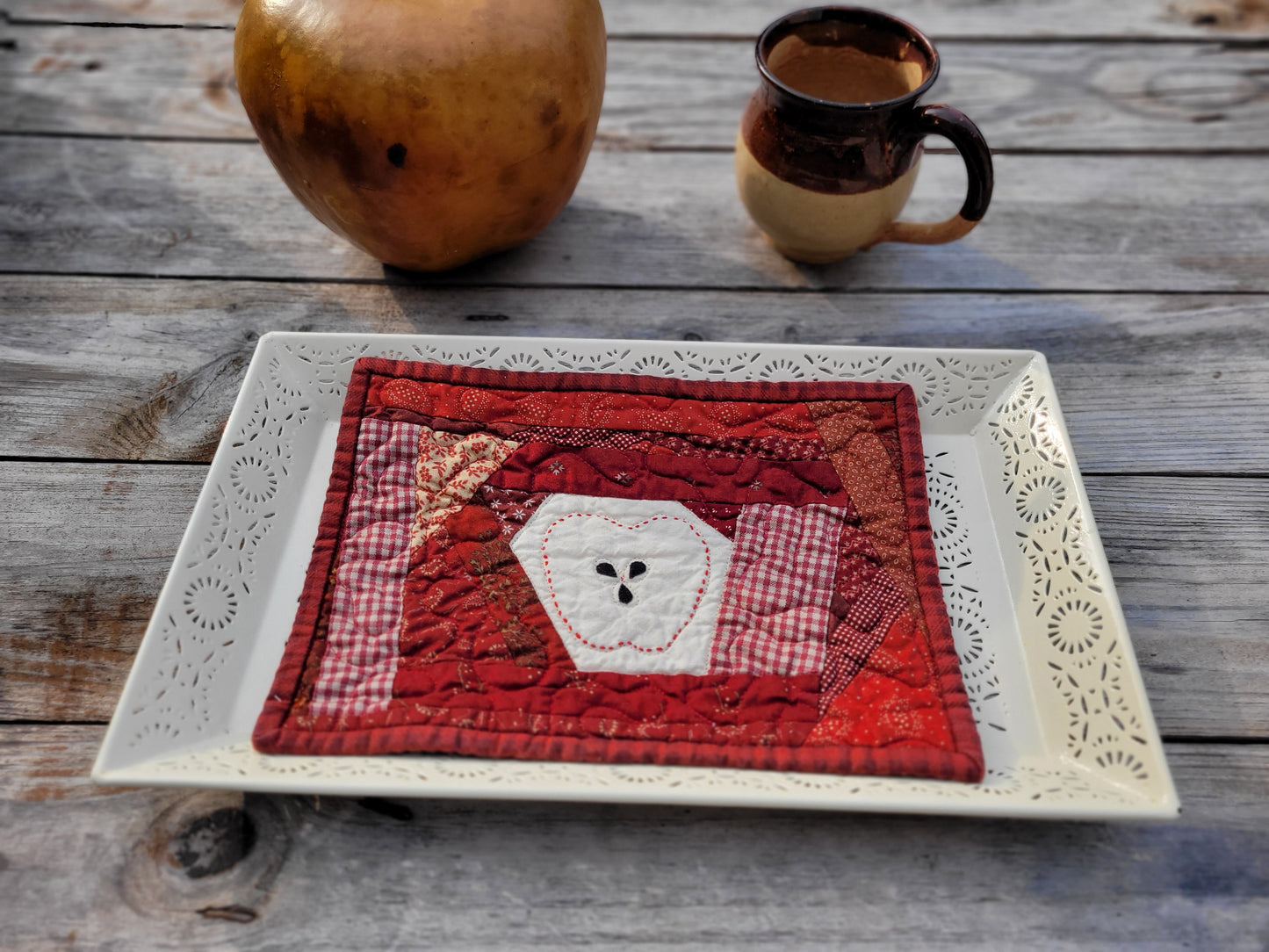 A handmade scrappy red patchwork quilted coaster with a central white embroidered apple shape, displayed on a white decorative platter on a rustic wooden table with a gourd and pottery mug in the background.