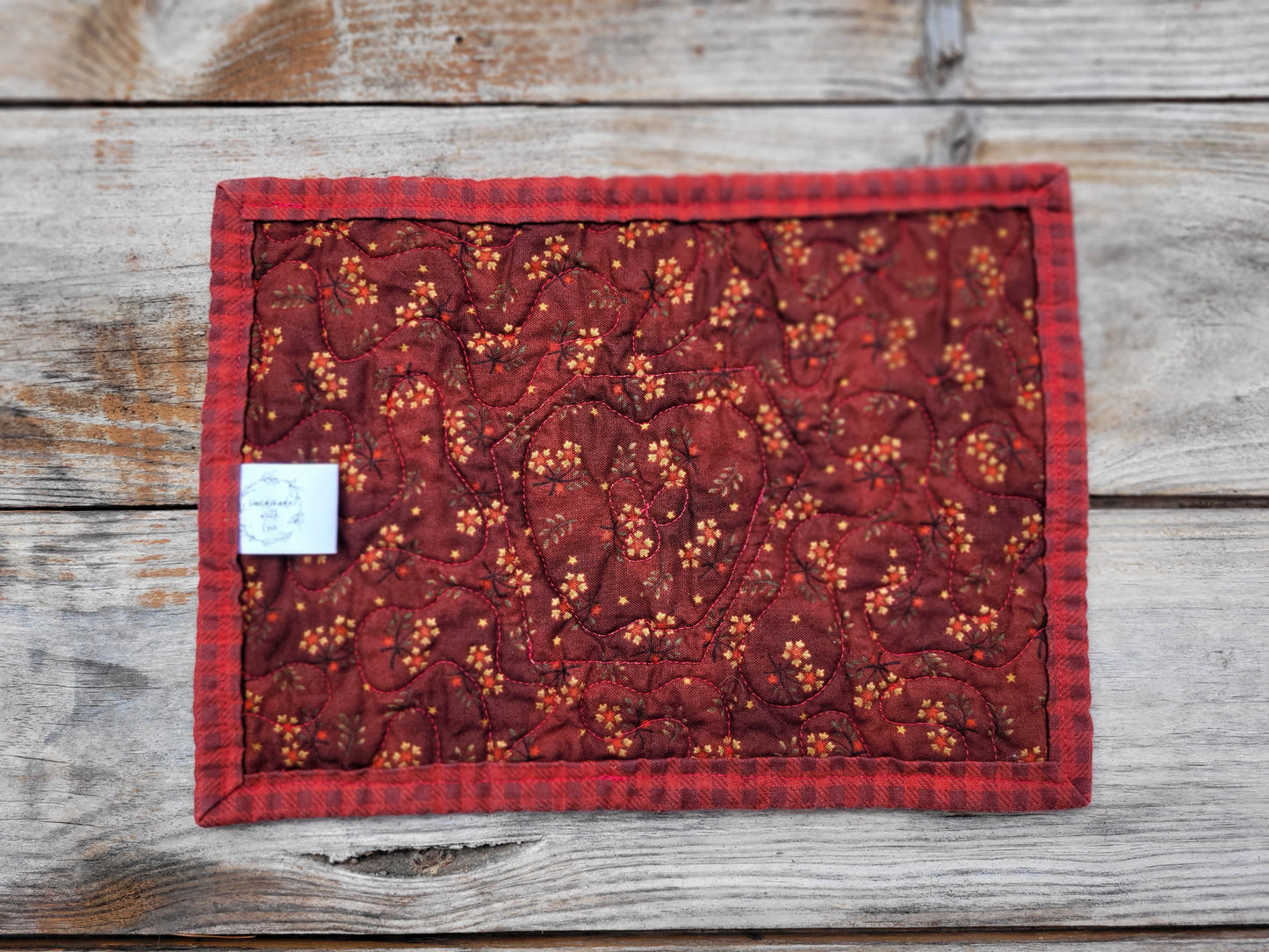 Showing the back of a rectangular quilted fabric coaster with a red and black buffalo plaid border and a red center featuring a floral vine pattern with small orange-yellow flowers, laid flat on a rustic gray wooden surface.