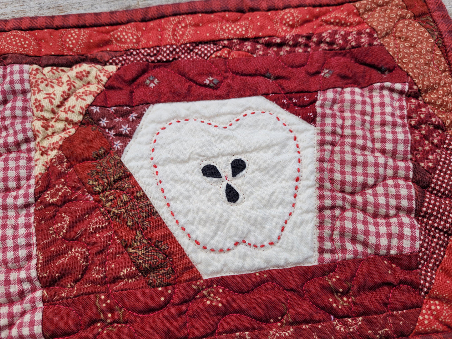 A close-up of a red and white quilted patchwork coaster featuring a central white patch with a stitched apple outline around three black seed details.