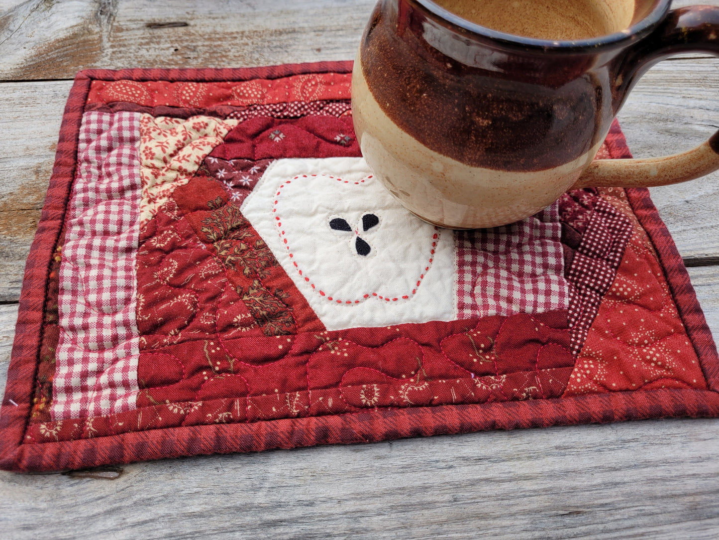 A handmade quilted patchwork mug rug in red and white tones, featuring a stitched apple design in the center, shown on a weathered wooden table with a pottery mug resting on top.