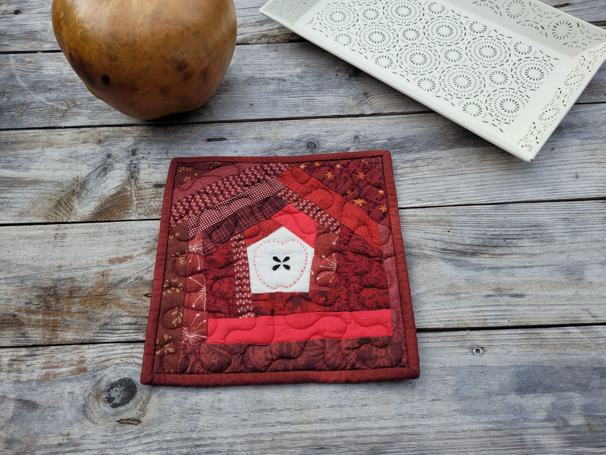 A square red quilted potholder featuring a white center that has a handstitched apple motif with black appliqued apple seeds, displayed on a rustic gray wooden tabletop with a decorative white tray and a brown gourd in the background.