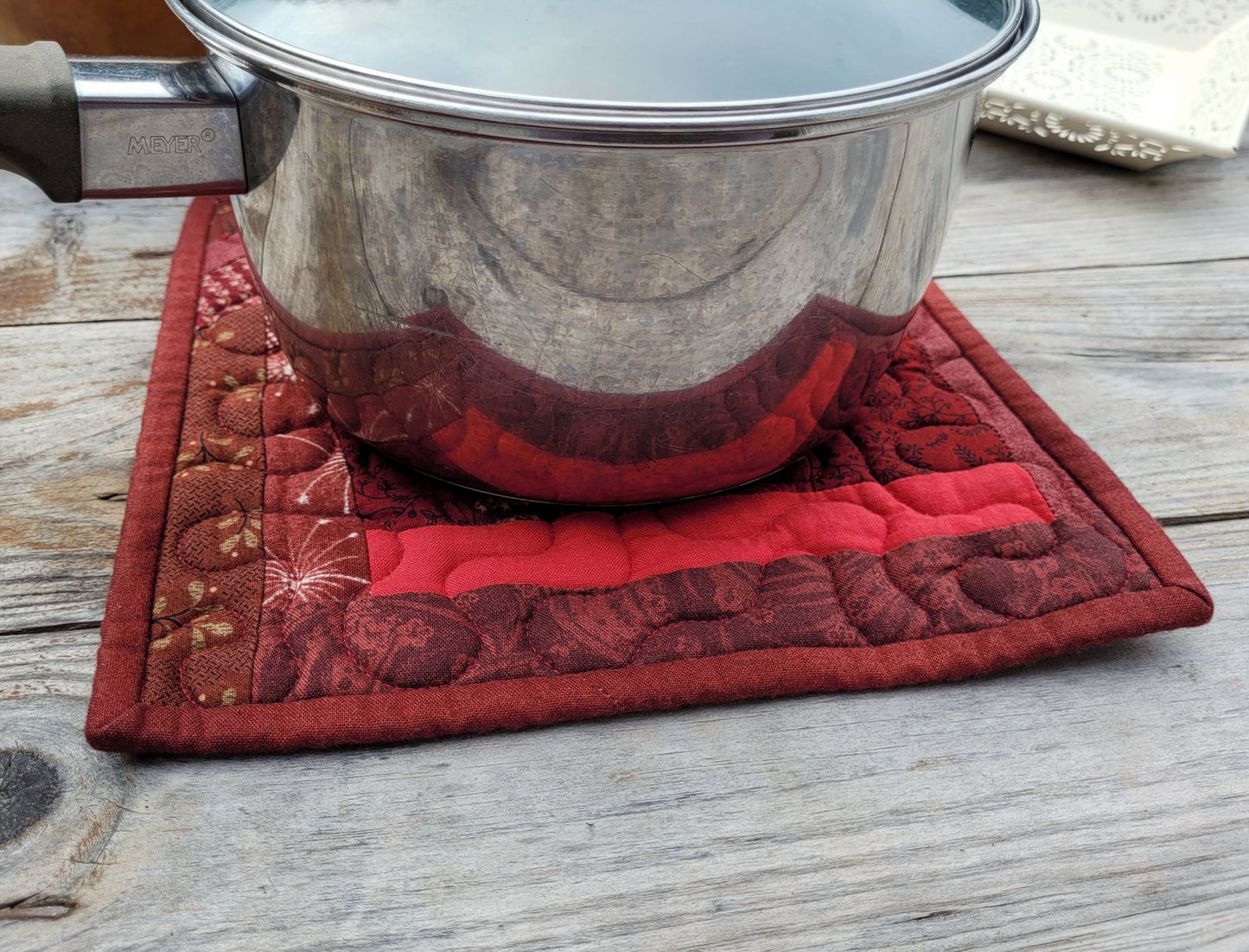 A square quilted red pot holder trivet with a floral pattern, supporting a stainless steel pot on a weathered gray wooden table.
