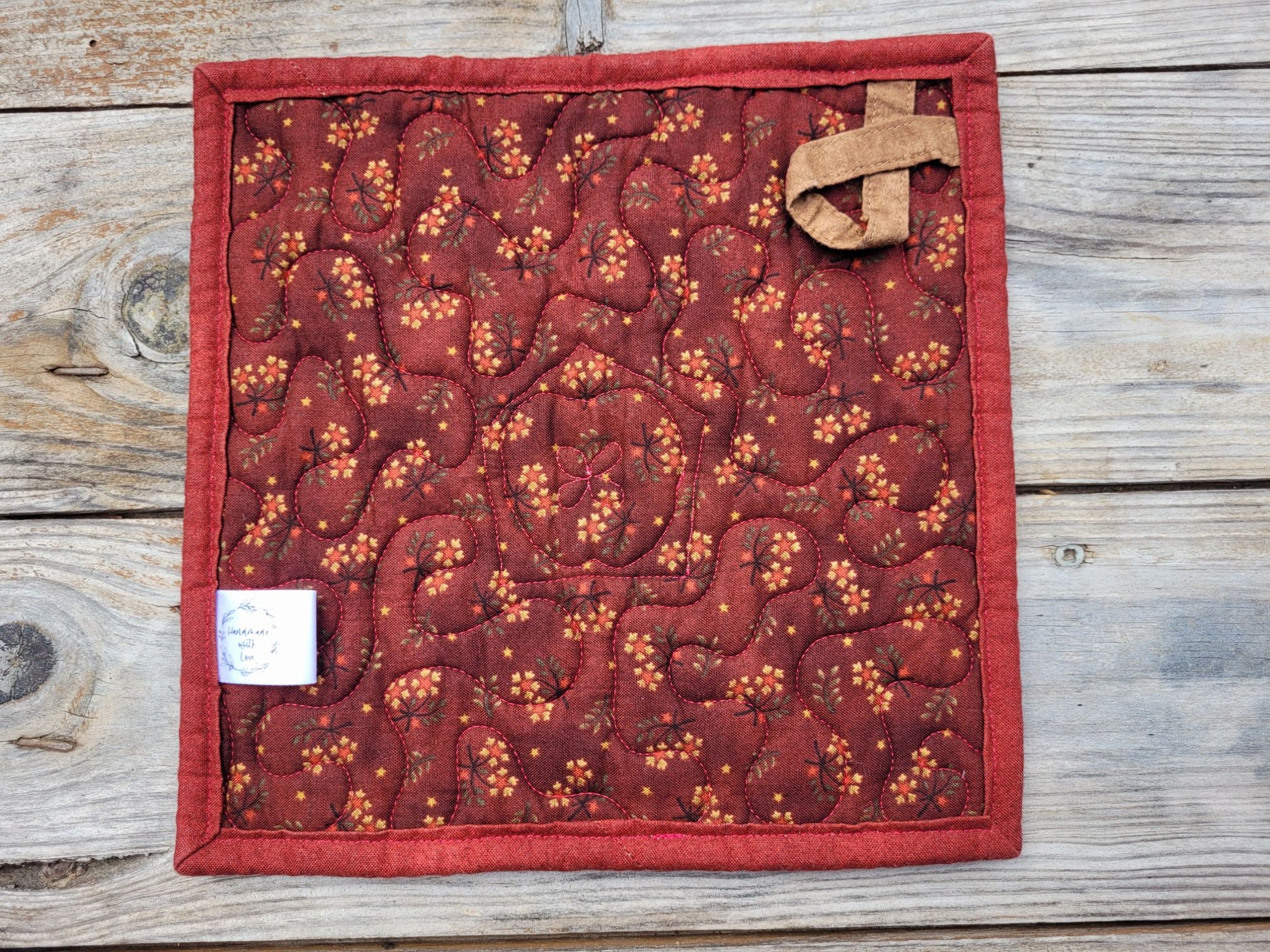 Showing the back of a square quilted fabric potholder or trivet in dark red with an orange floral pattern and a tan fabric hanging loop, shown on a weathered wood plank background.