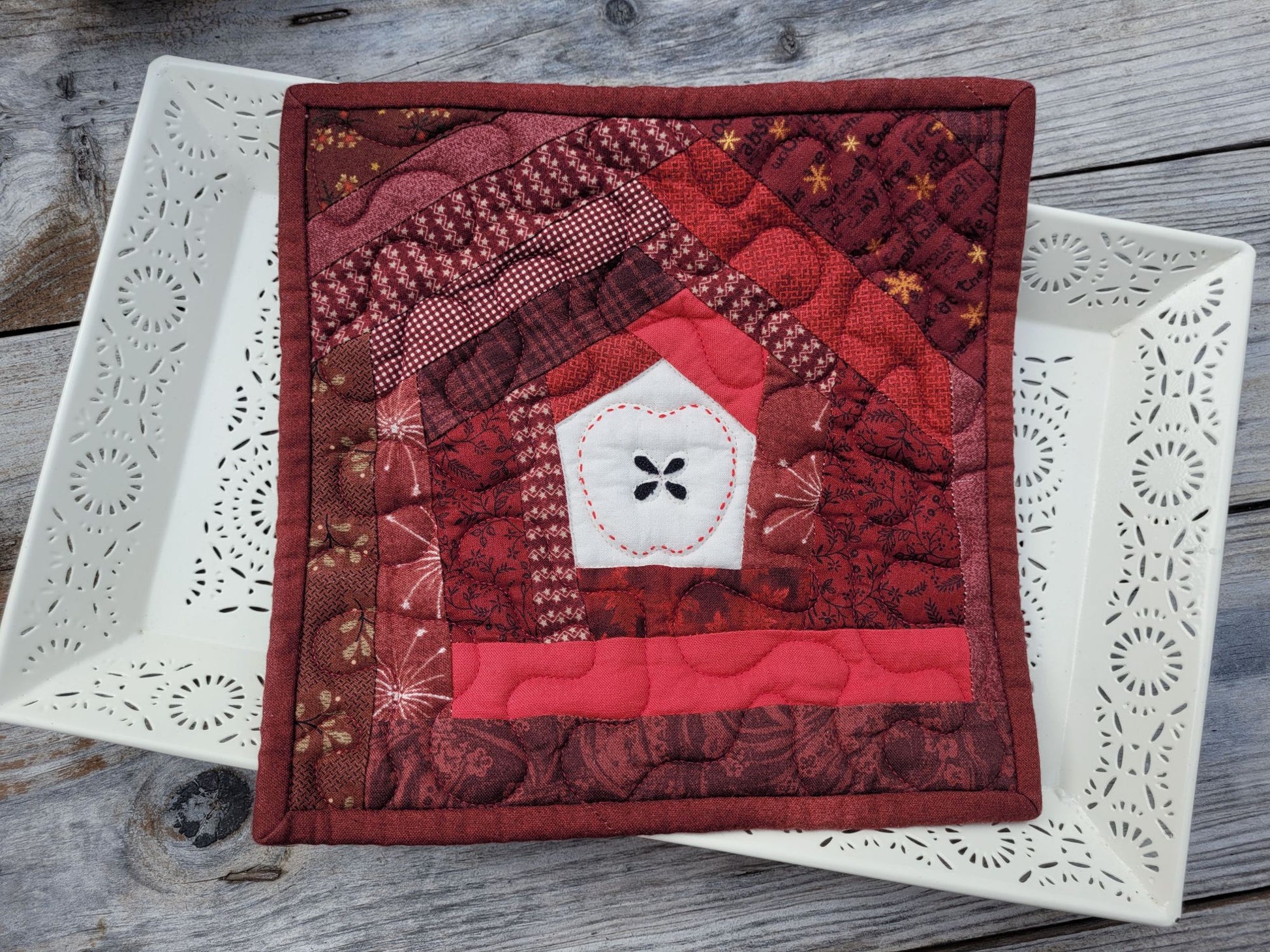 A square quilted patchwork hot mat featuring a handstitched apple design in the centre. Scrappy patchwork is in shades of red, burgundy, and white, displayed on a white ceramic tray over a rustic gray wooden surface.