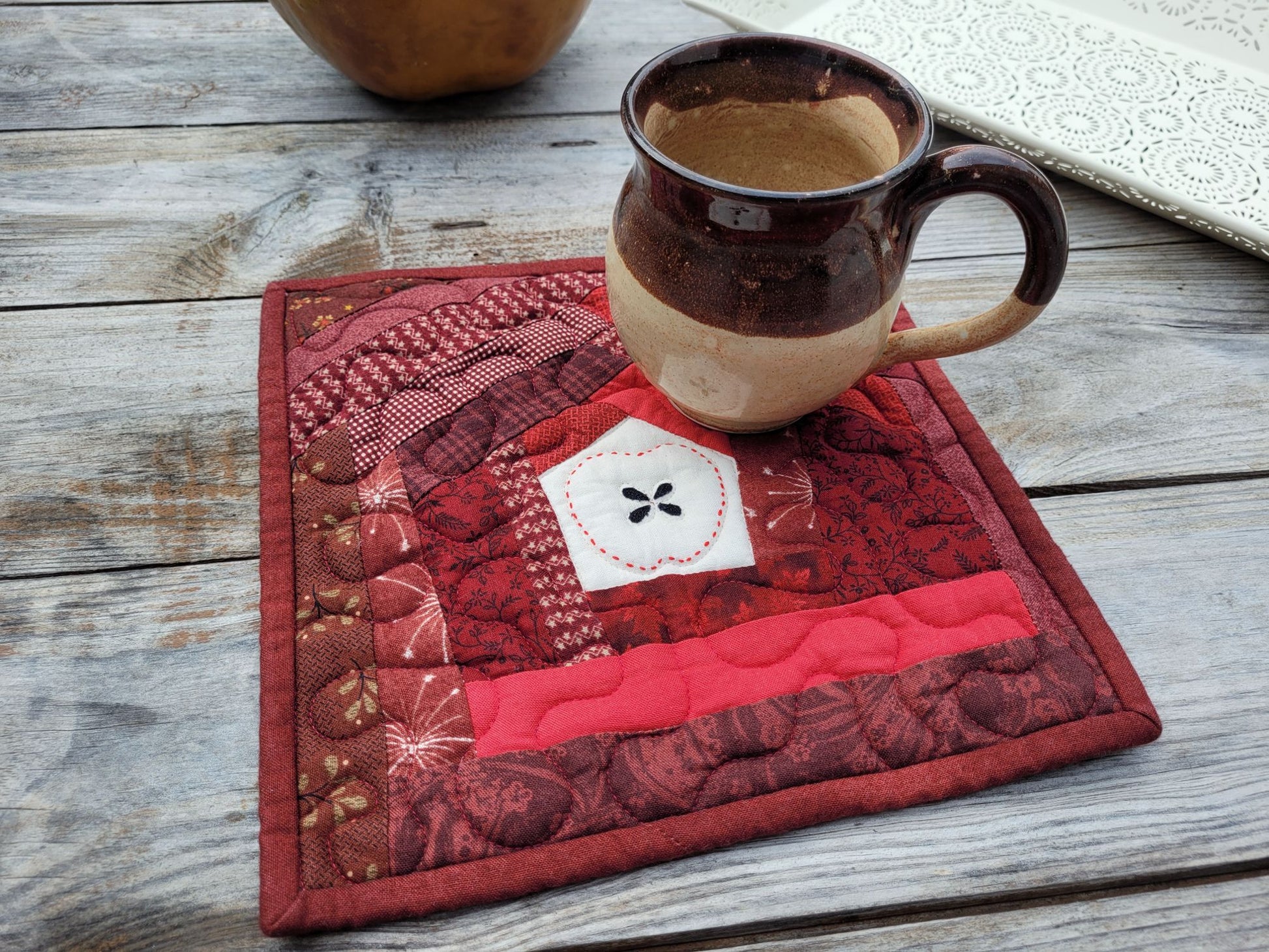 A square quilted apple themed potholder in shades of red, featuring a small white muslin patch in the centre with a handstitched apple outline around appliqued seeds, shown on a rustic wooden table with a pottery mug resting on top.