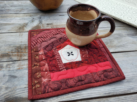 A square quilted apple themed potholder in shades of red, featuring a small white muslin patch in the centre with a handstitched apple outline around appliqued seeds, shown on a rustic wooden table with a pottery mug resting on top.