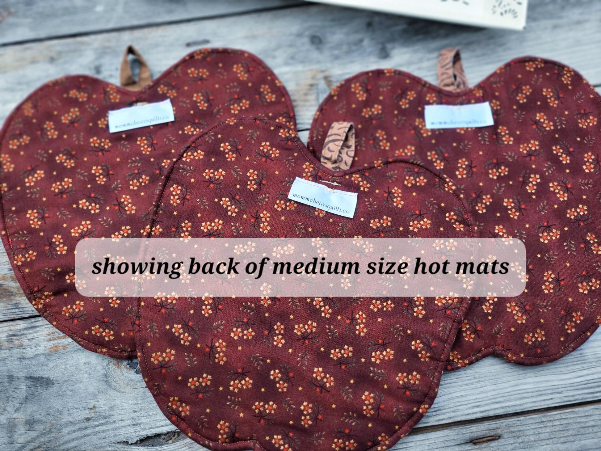 Three apple-shaped hot mats with a deep red and orange floral pattern are arranged on a weathered gray wooden surface. A text overlay reads 'showing back of medium size hot mats'.