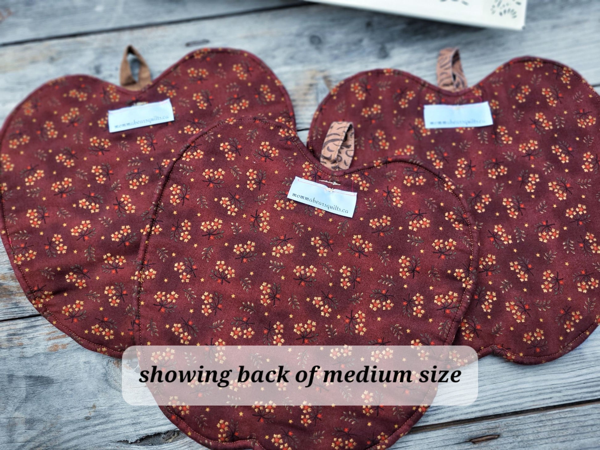 Three small apple-shaped quilts with a dark red and orange floral pattern are arranged on a weathered gray wooden surface. A text overlay at the bottom reads 'showing back of medium size'.