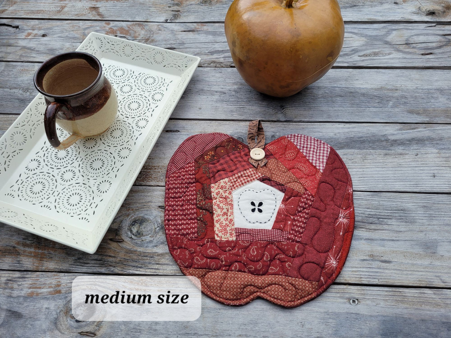 A small apple shaped quilt displayed on a rustic wooden table with a white decorative tray, a pottery mug and a rustic golden apple shaped gourd.