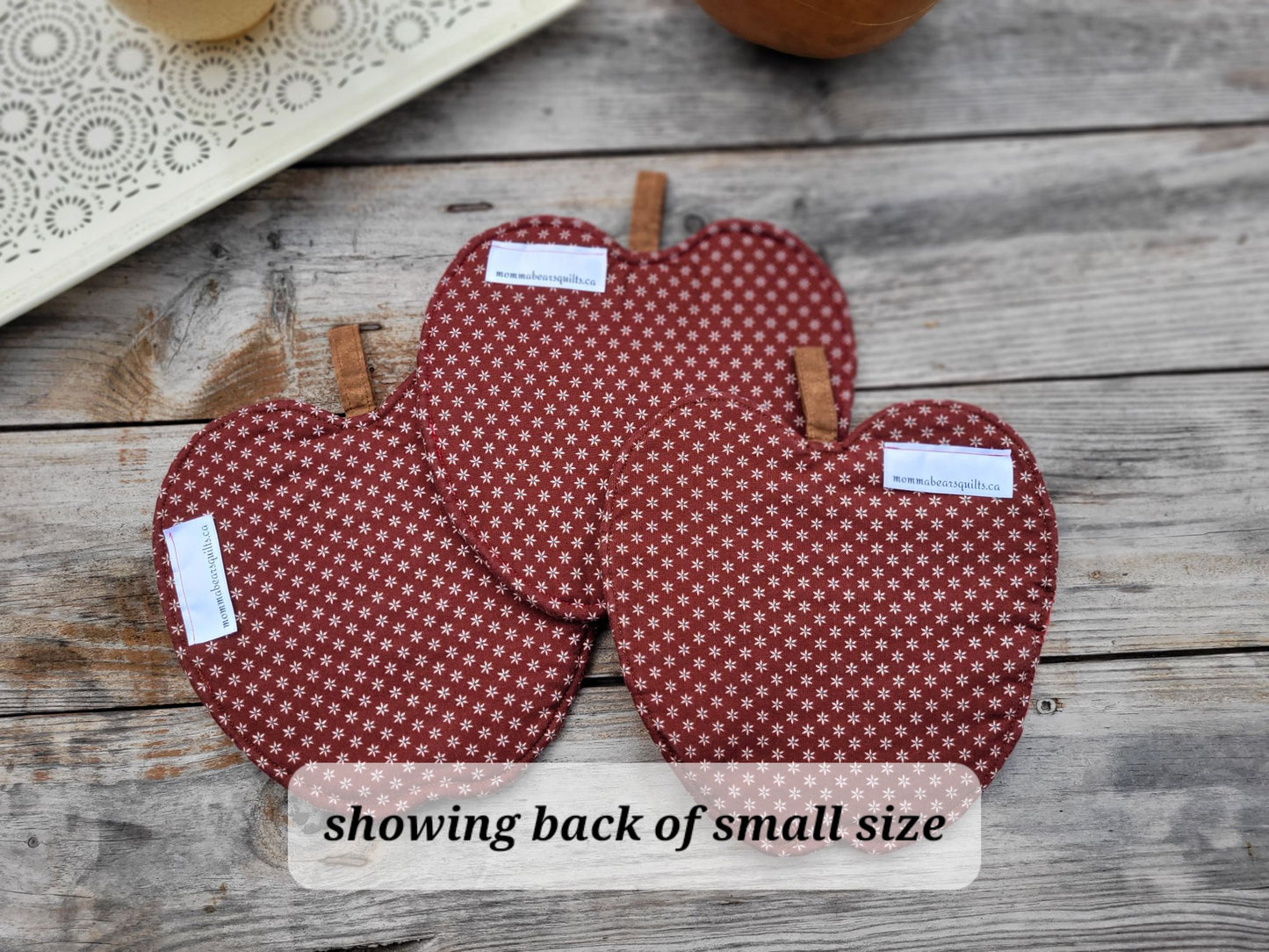 Three apple-shaped fabric potholders with a red and white floral pattern are arranged on a rustic wooden surface. A text overlay at the bottom reads 'showing back of small size'.