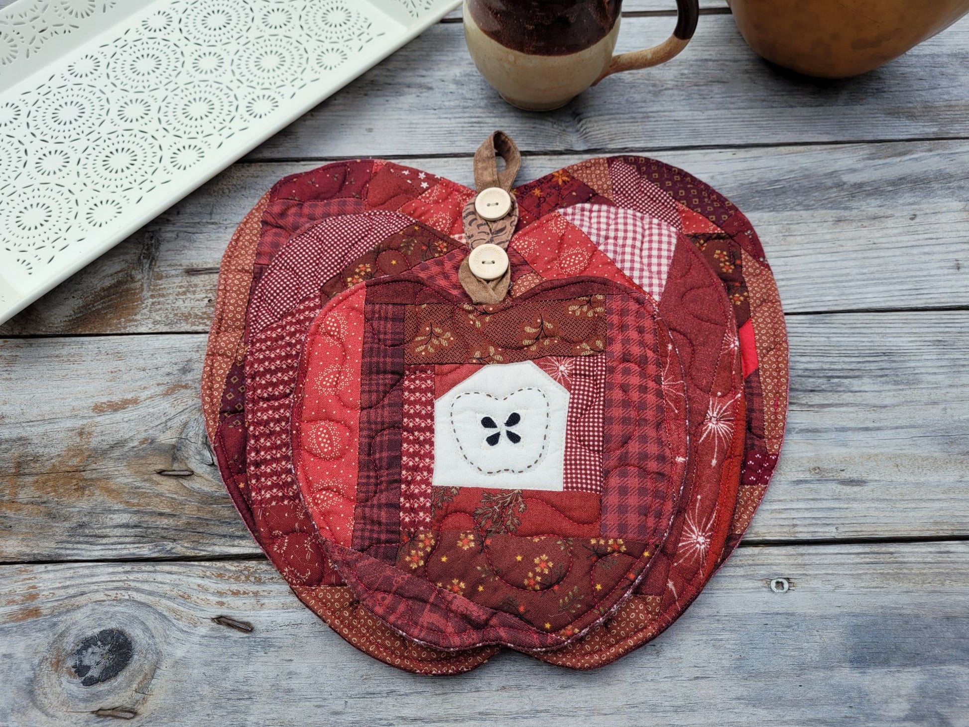 A set of three apple-shaped patchwork quilts are nested together into a gift set. Apples feature scrappy patchwork  in shades of red, burgundy, and brown, featuring a central white handstitched apple motif. Wooden buttons secure the hanging loops on two of the mats and allow one to hang in front of the other decreasing in size. They are displayed on a rustic gray wooden table wth a white decorative tray partially visable.