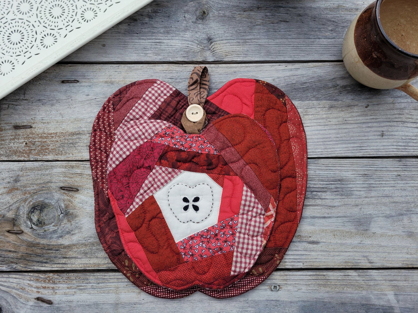 A red and pink apple-shaped quilted fabric potholder with a stitched apple core and a wooden button handle, displayed on a rustic gray wooden tabletop next to a ceramic mug.