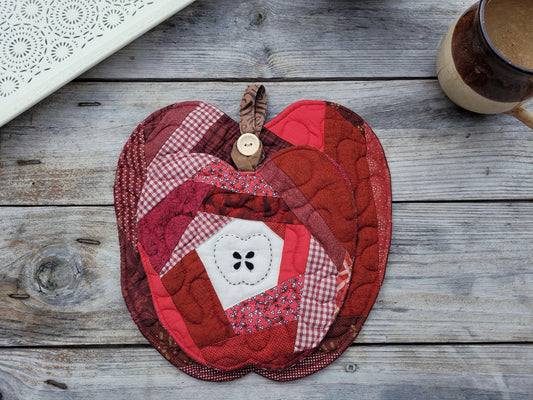 A red and pink apple-shaped quilted fabric potholder with a stitched apple core and a wooden button handle, displayed on a rustic gray wooden tabletop next to a ceramic mug.