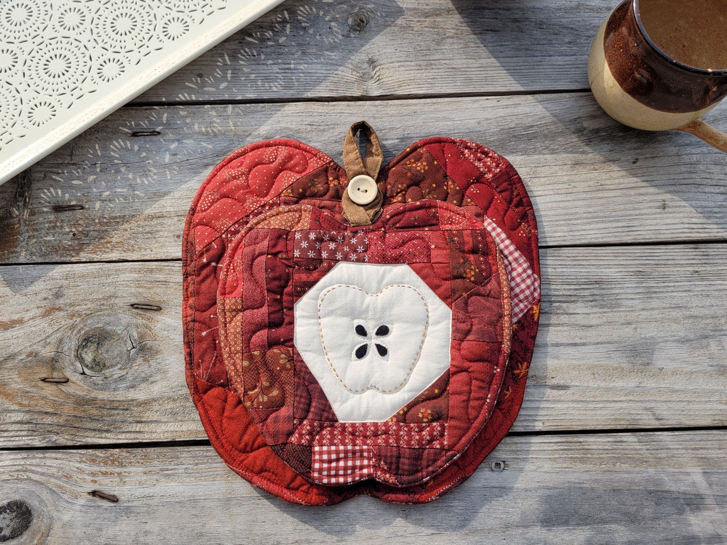 Two small apple-shaped mini quilts in red and brown fabrics with a white apple core design are nested together using the button hanging loop on the larger one to hang the smaller one in front as an appealing gift set. They are displayed on a rustic wooden surface.
