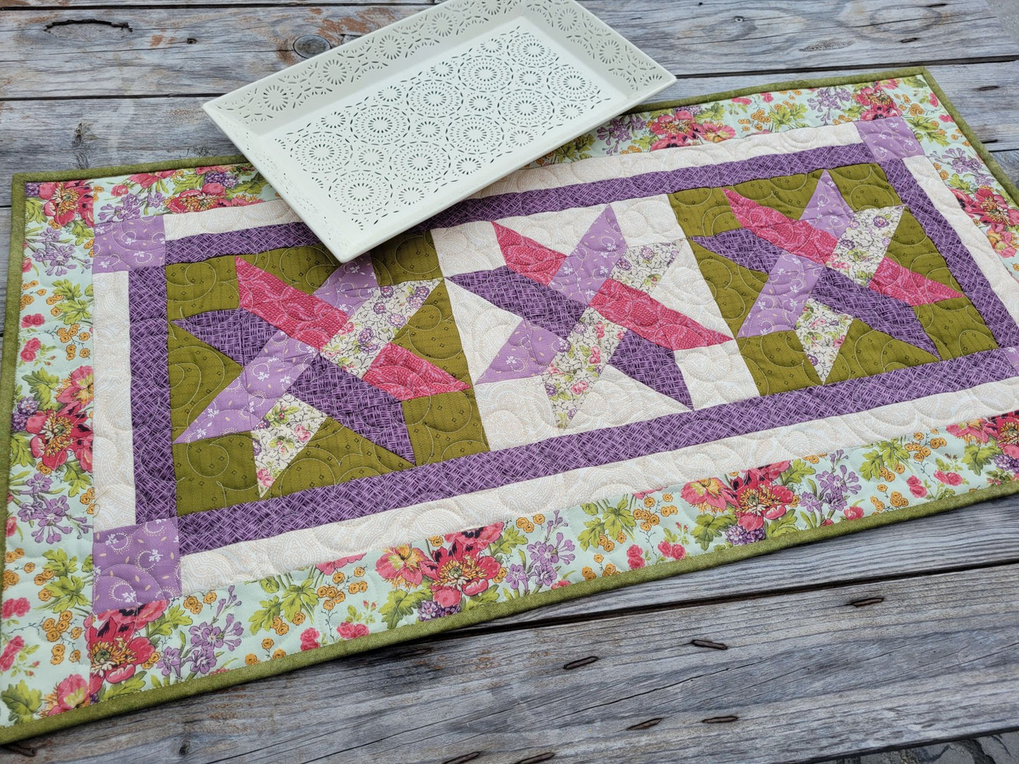 A handmade quilted table runner with a floral border and three pink and purple woven star patterns, styled on a rustic gray wooden table with a white decorative serving tray.