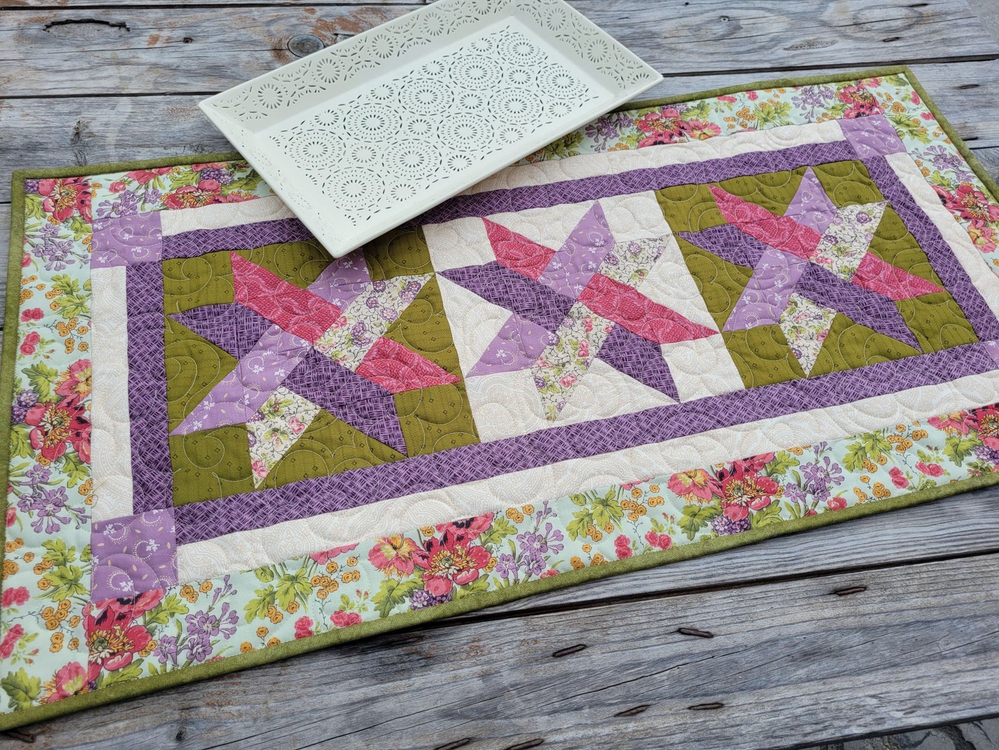 A handmade quilted table runner with a floral border and three pink and purple woven star patterns, styled on a rustic gray wooden table with a white decorative serving tray.