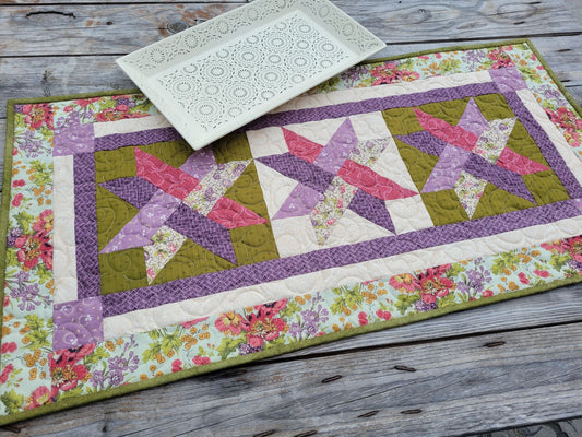 A handmade quilted table runner with a floral border and three pink and purple woven star patterns, styled on a rustic gray wooden table with a white decorative serving tray.