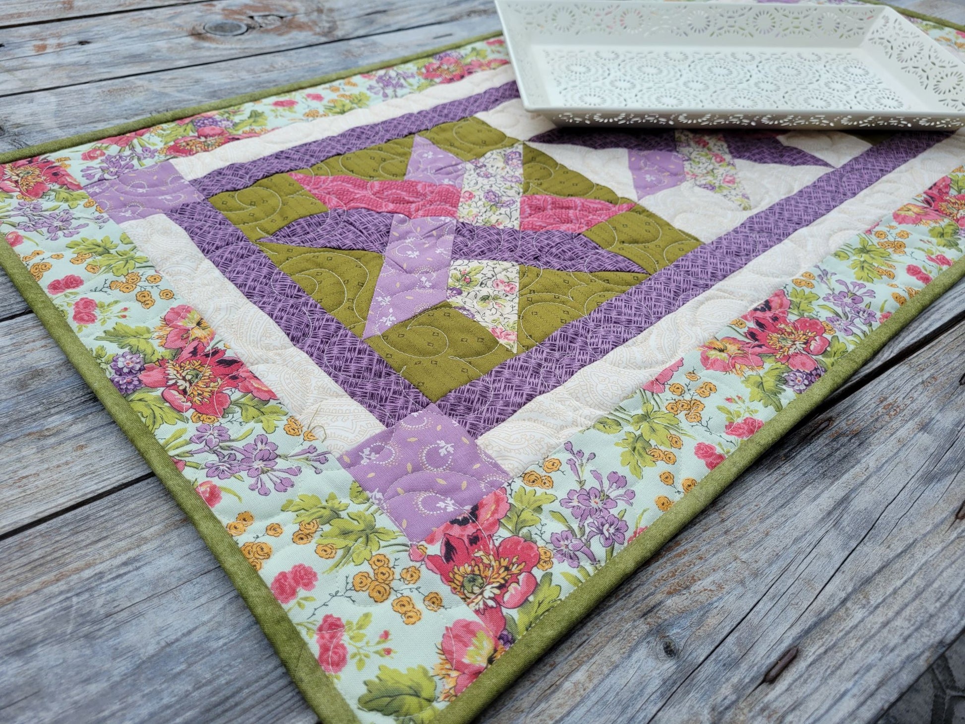 A handmade quilted table runner with a floral border and a central star patchwork design in purple, moss green, and pink, displayed on a rustic wooden table with a white decorative tray.
