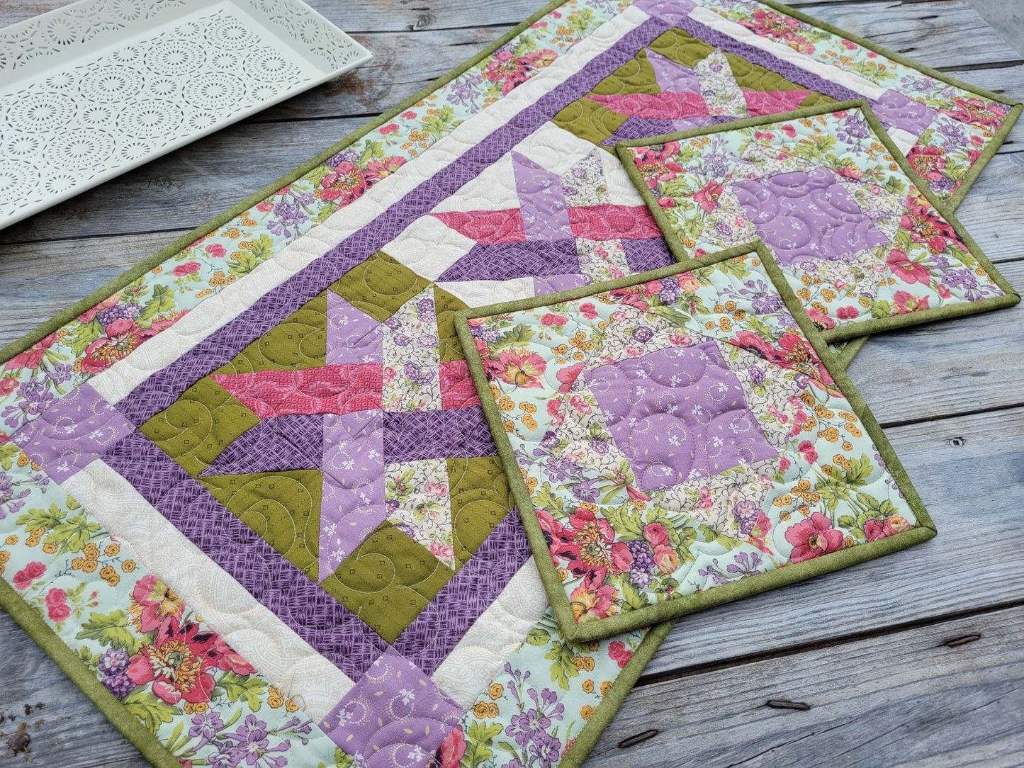 A quilted table runner and matching square hot mats laid out on a rustic gray wooden surface. The quilt features a patchwork design with purple, pink, and moss green floral and geometric patterns.