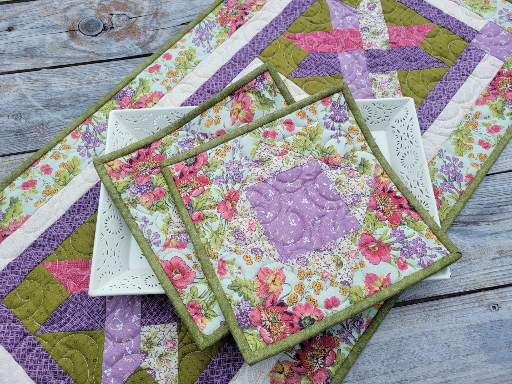 Two square quilted floral potholders with moss green binding are arranged on a white decorative tray sitting atop a matching quilted table runner on a rustic gray wooden tabletop.