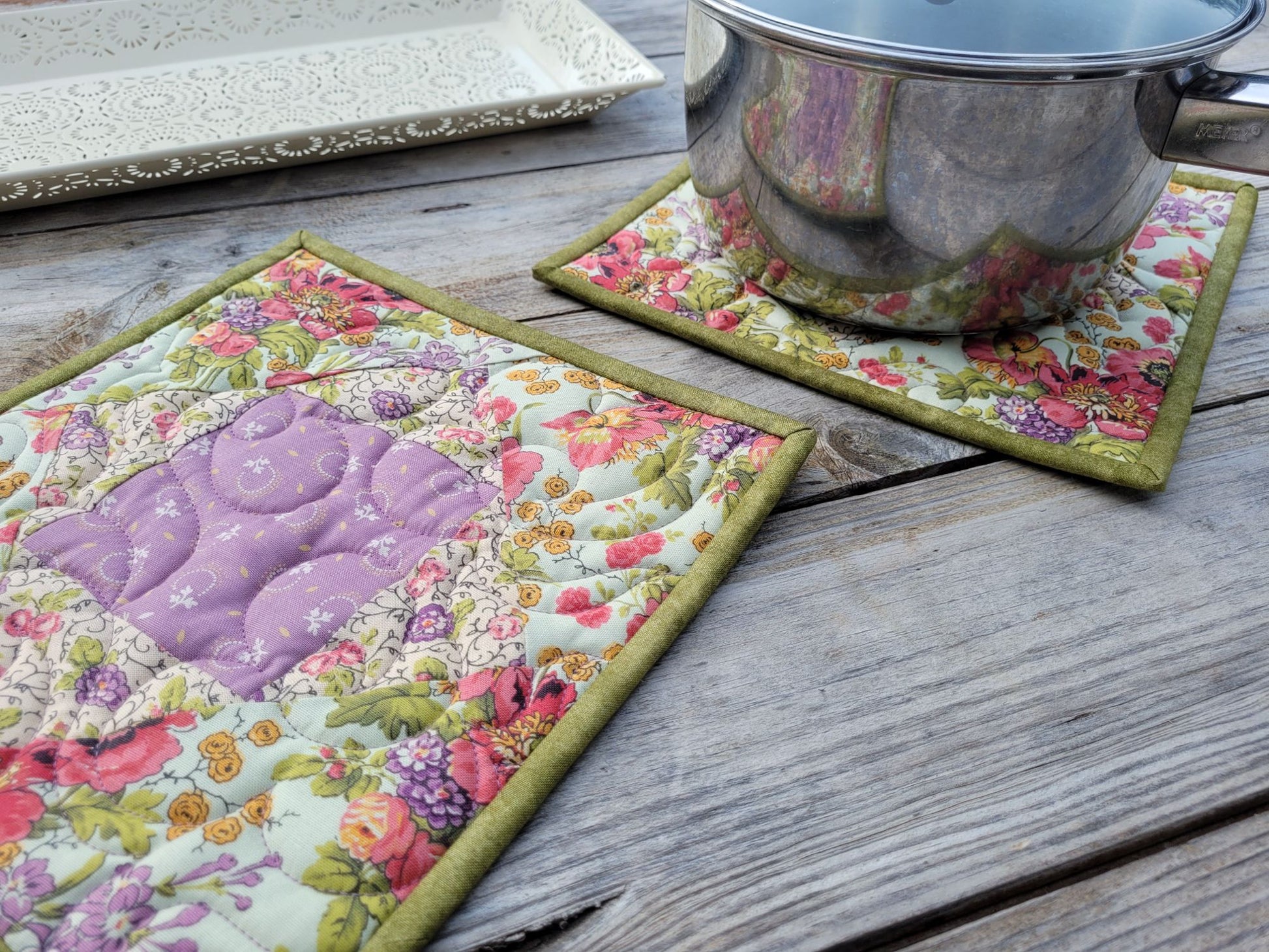 Two floral quilted potholders with moss green binding sit on a weathered wooden table, with a medium size saucepan resting on one and a decorative white tray partially visible in the background.