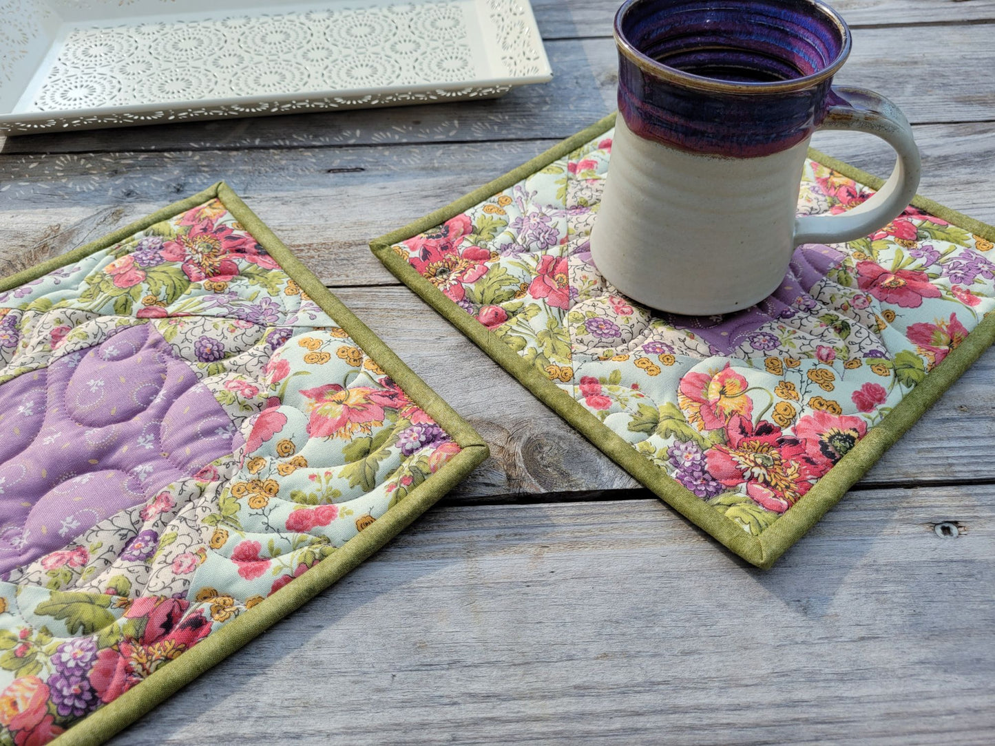 Two handmade quilted floral potholders on a weathered gray wooden table, with a ceramic mug resting on one. A white decorative tray is in the background.