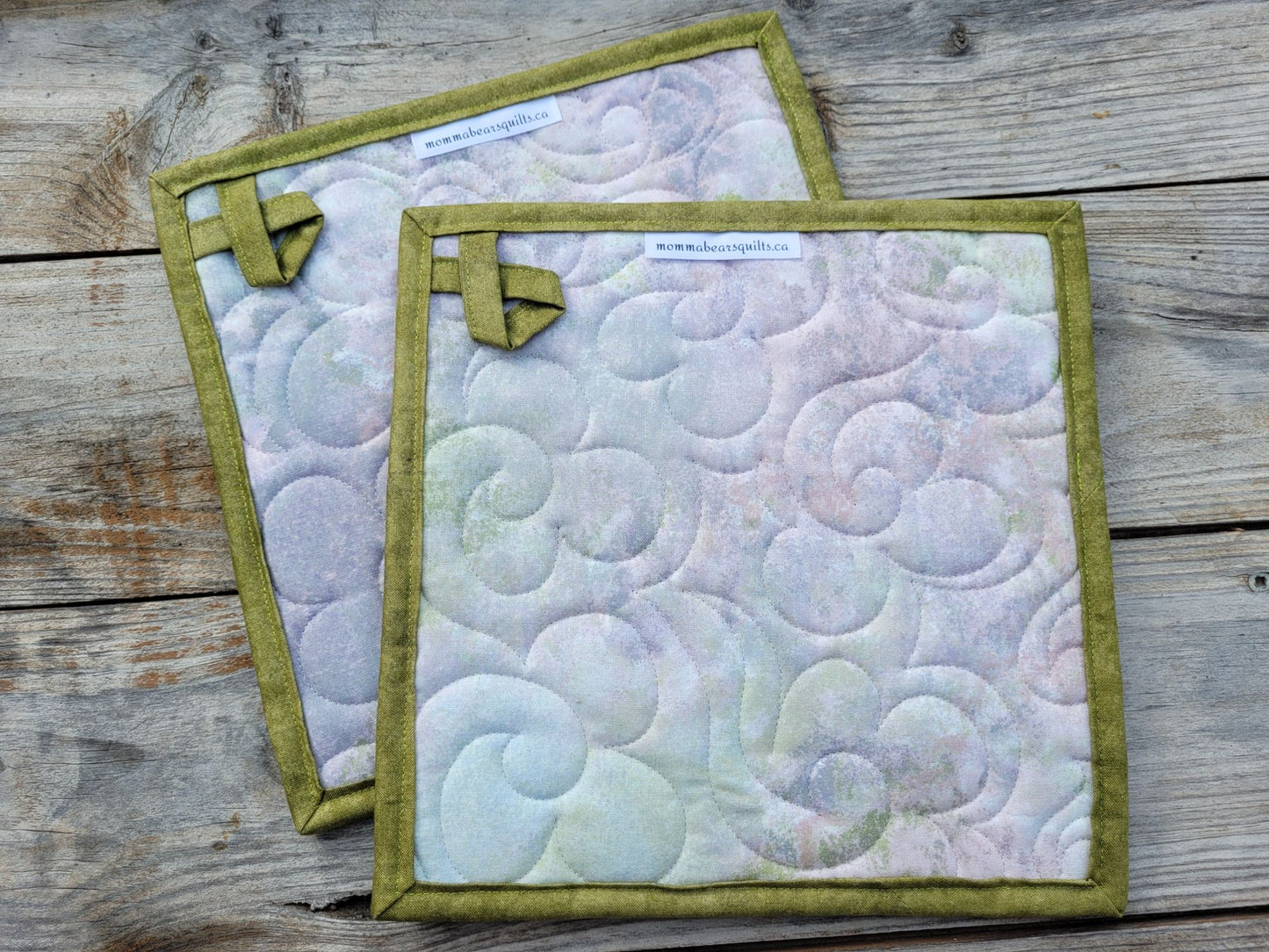 Showing the back of two quilted fabric potholders with moss green borders and hanging loops, featuring a pastel swirling pattern in lavender, blue, and green, displayed on a rustic wooden surface.