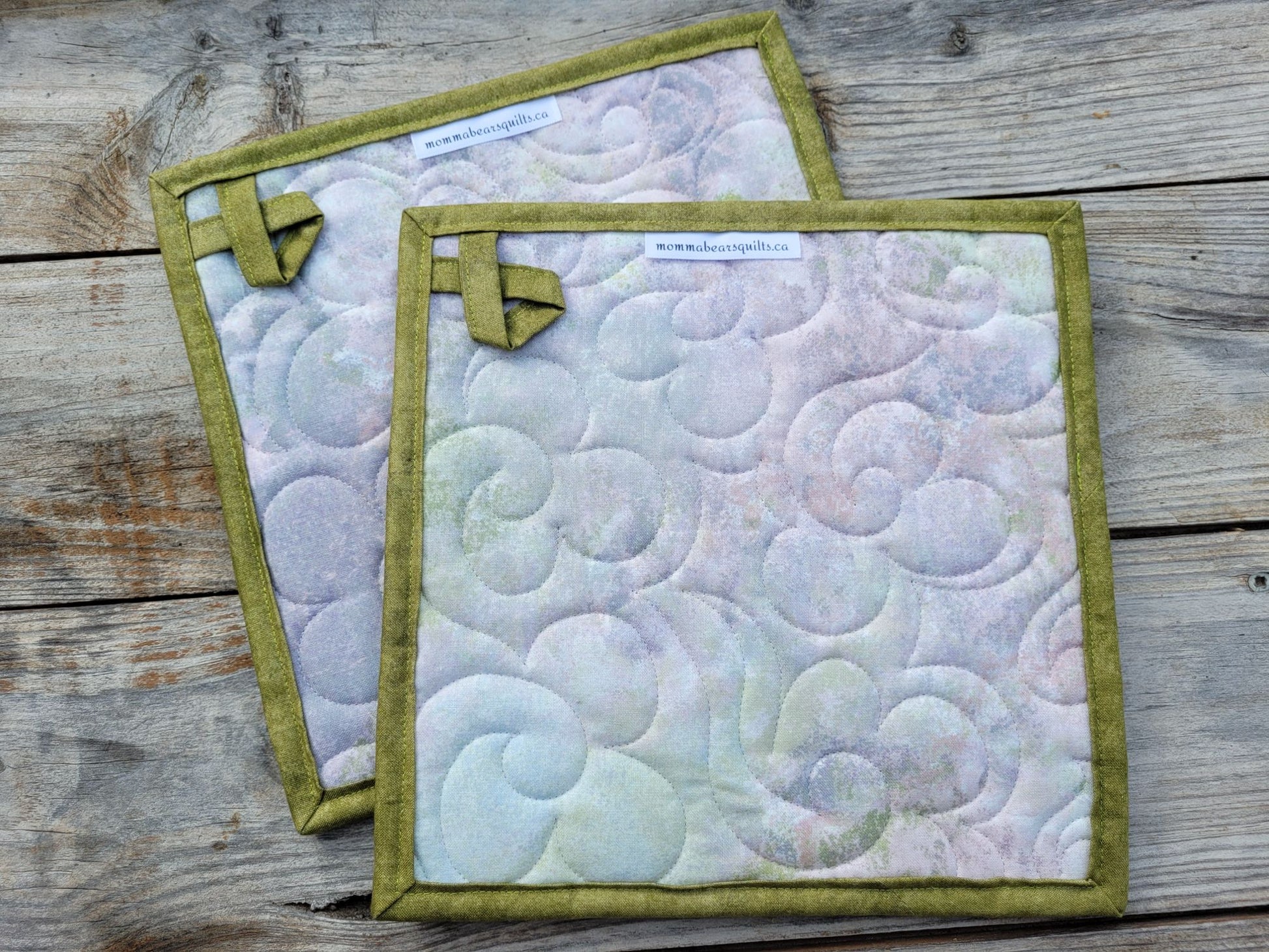 Showing the back of two quilted fabric potholders with moss green borders and hanging loops, featuring a pastel swirling pattern in lavender, blue, and green, displayed on a rustic wooden surface.
