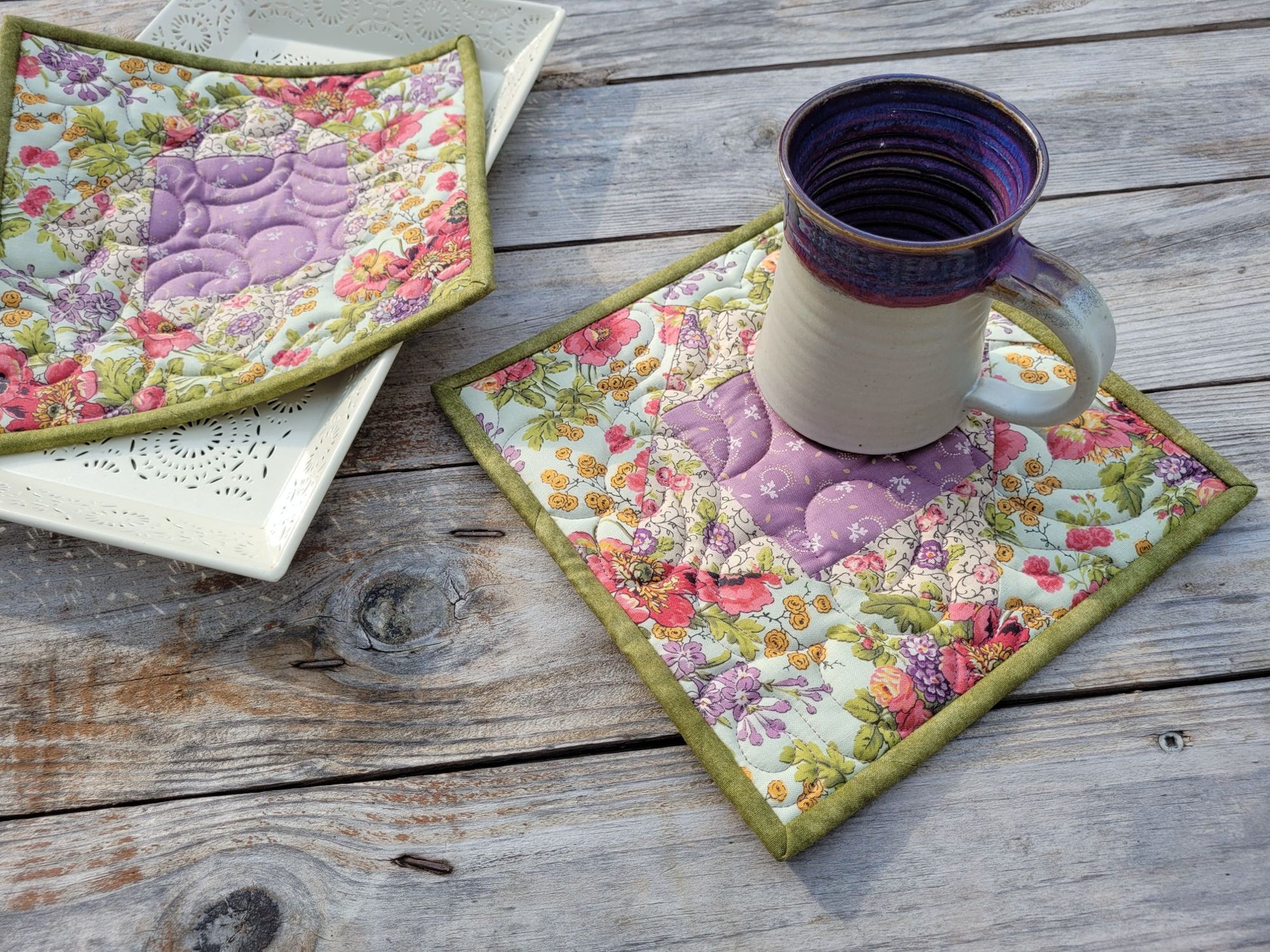 Two quilted floral hot mats with purple floral patchwork motifs are displayed on a rustic wooden table. One hot mat is shown flat with a pottery mug, and the other is angled on top of a white decorative tray.