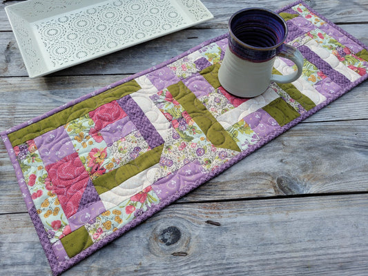 A rectangular quilted table runner with purple, pink, and moss green floral patchwork, styled on a rustic gray wooden table with a pottery mug and a white decorative tray.