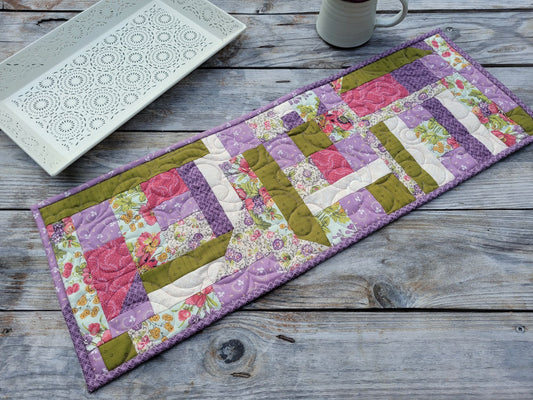 A handmade quilted table runner with a patchwork design featuring purple, pink, and moss green fabrics, displayed on a rustic gray wooden table next to a white ceramic tray and a mug.
