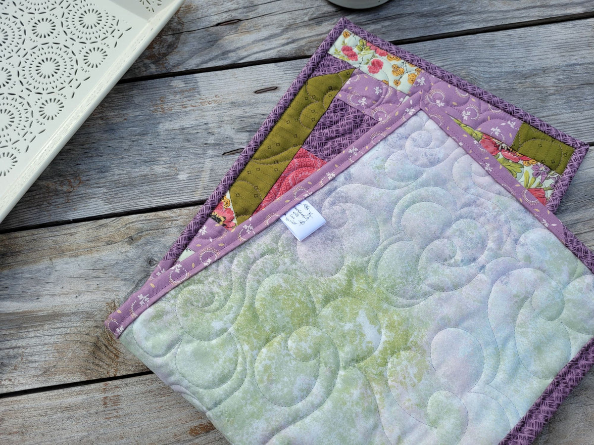 A close-up of a handmade quilted table runner folded to show the backing in a mottled fabric in tones of pastel colours of green, lavender, pink and blue. Handmade label tag is shown on side of runner. Styled on a rustic wooden table with a partially visable white decorative tray.