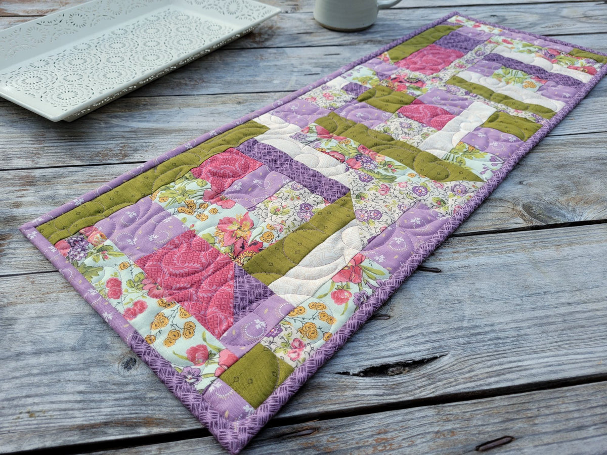 Angle view of a handmade quilted table runner styled on a rustic wooden table with a decorative tray in the background.