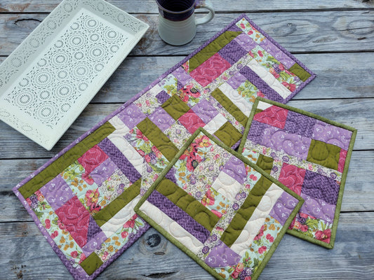 Three handmade patchwork crazy quilts in purple, pink, and moss  green are arranged on a weathered gray wooden table, styled with a white decorative tray and a mug. There is one small size table runner with purple binding laying partially beneath  two hotmats sitting, which have moss green binding. All coordinating fabrics.