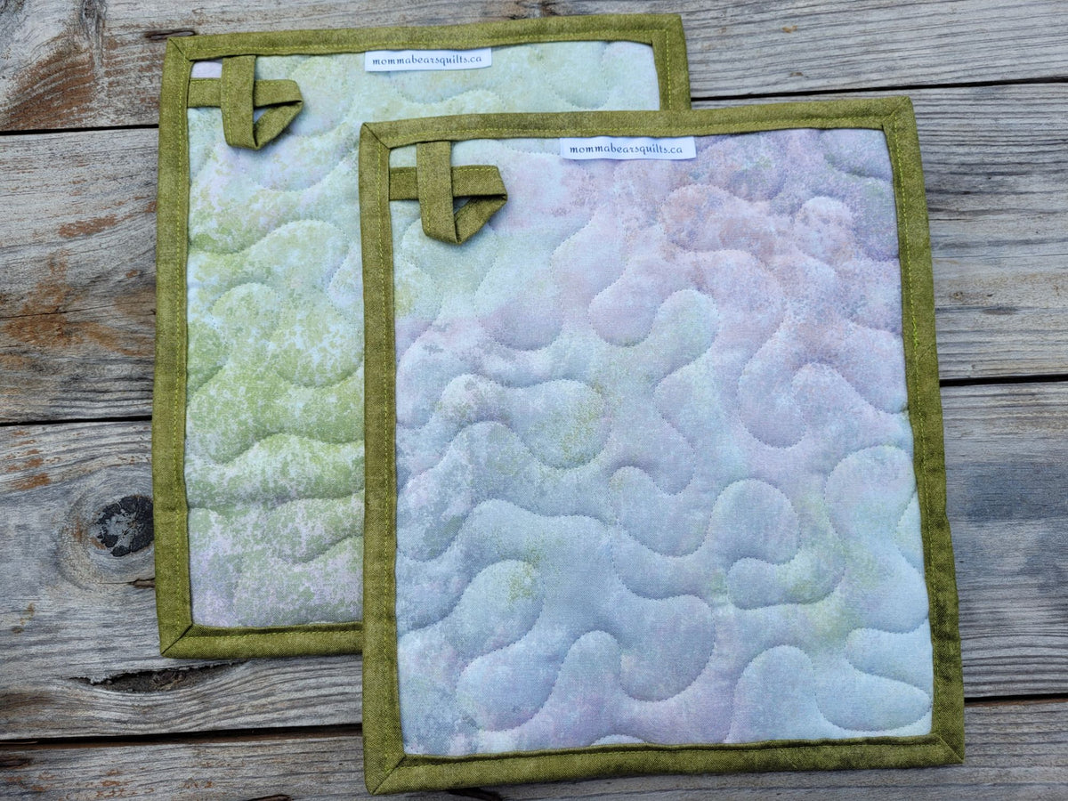 Showing the back of two quilted fabric potholders with moss green binding and hanging loops, displayed on a rustic wooden surface. Backing is a mottled fabric in pastel shades of green, lavender, pink, and white so that the colour placement is different  on each hot mat.
