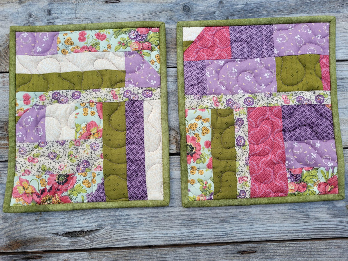 Two handmade quilted fabric hot mats with a crazy quilt patchwork design in purple, pink, moss green, and floral prints, displayed side-by-side on a weathered gray wooden surface.