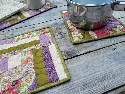 A close-up of two handmade quilted hot mats on a weathered wooden table, with one mat holding a medium size saucepan. Matching runner is partially visable in the top left with a white decorative tray and mug. 