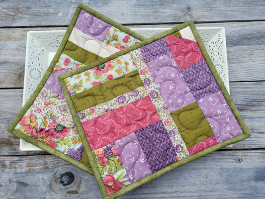 Two square quilted patchwork hot mats with moss green borders, displayed on a white decorative tray sitting on a rustic gray wooden table.