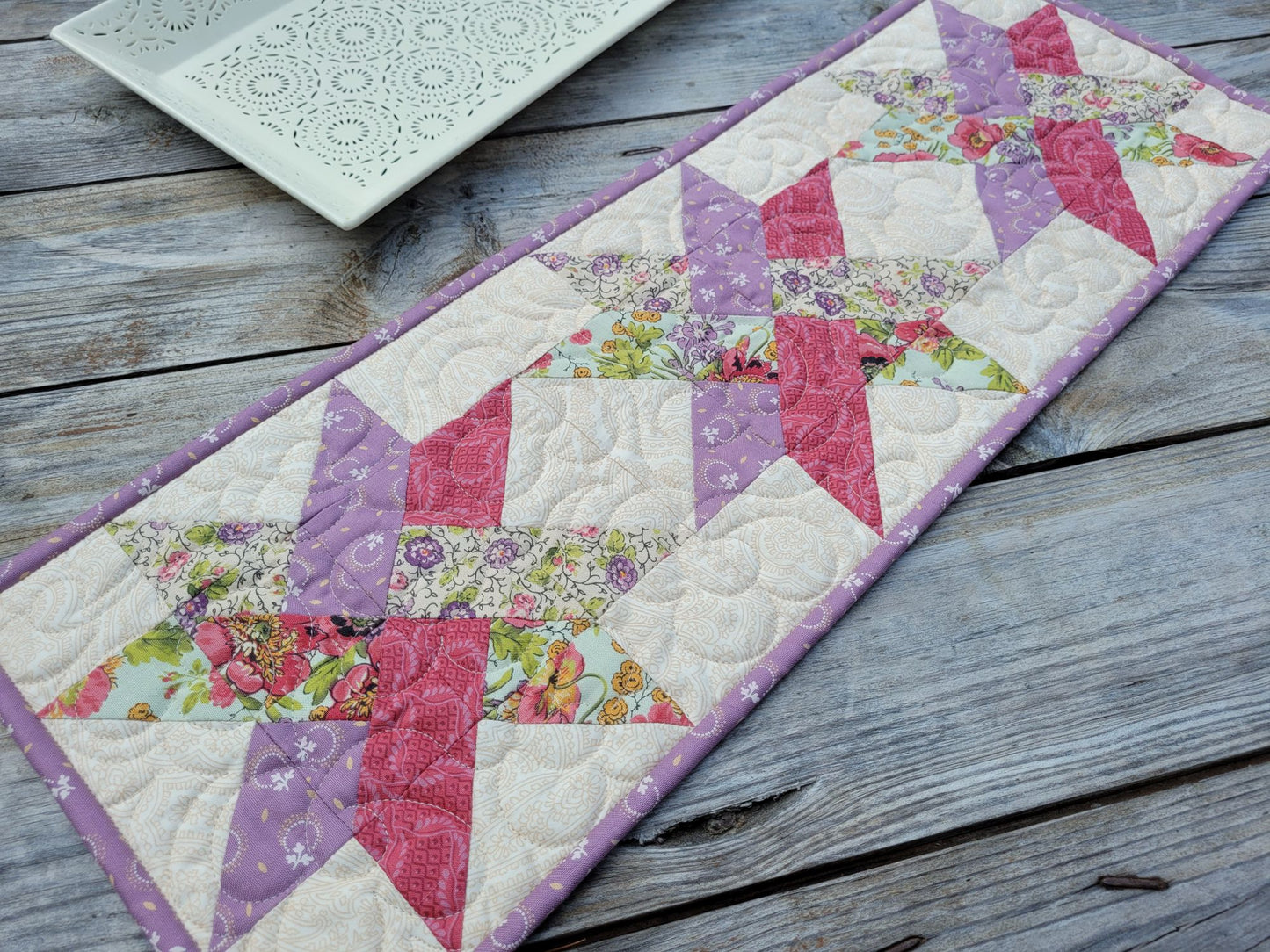 A handmade quilted table runner with a woven star pattern in pink, purple, and floral print fabrics, laid out on a rustic gray wooden table. A white decorative tray is partially visable in the background.
