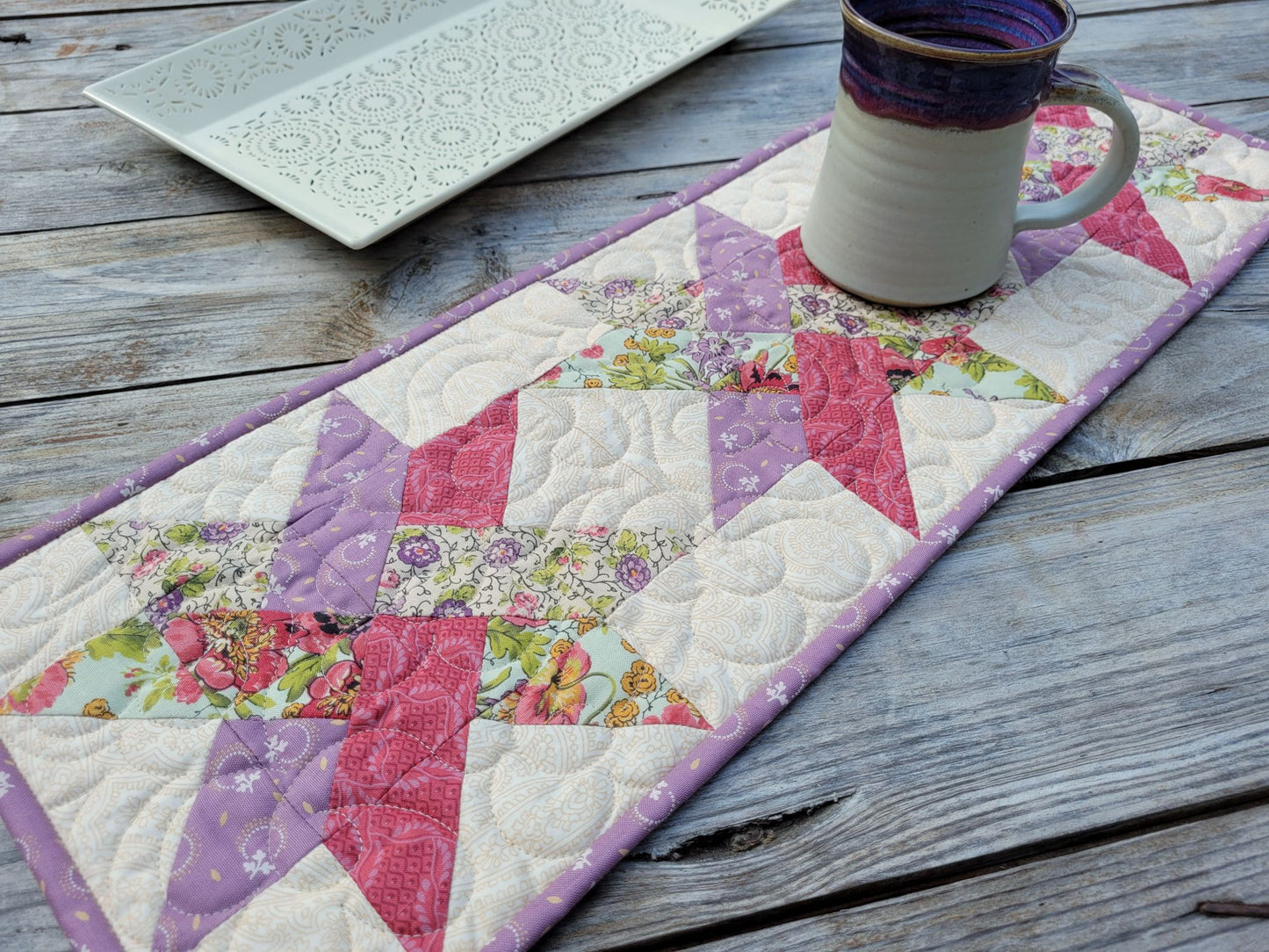A quilted star-pattern table runner in pink, purple, and floral print, styled on a rustic wooden table with a ceramic mug and a white decorative tray.