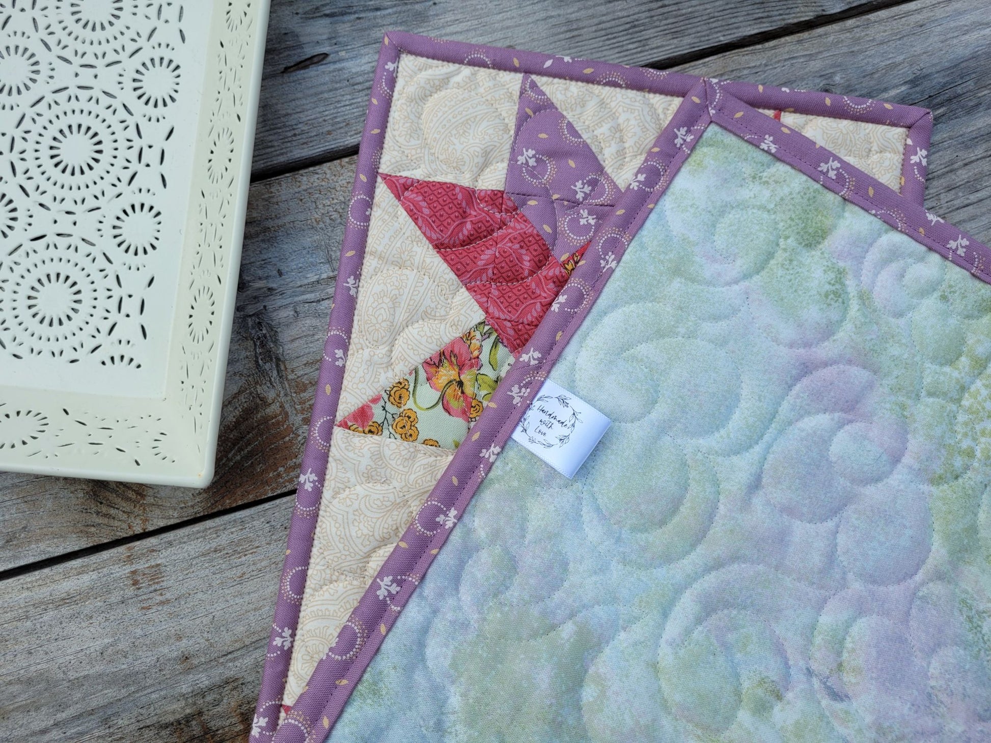 A close-up of a handmade quilted table runner folded to show the pastel green and lavender swirl patterned backing with purple borders and a glimpse of the woven star patchwork on the front. It is displayed on a rustic wooden surface next to a white decorative tray.