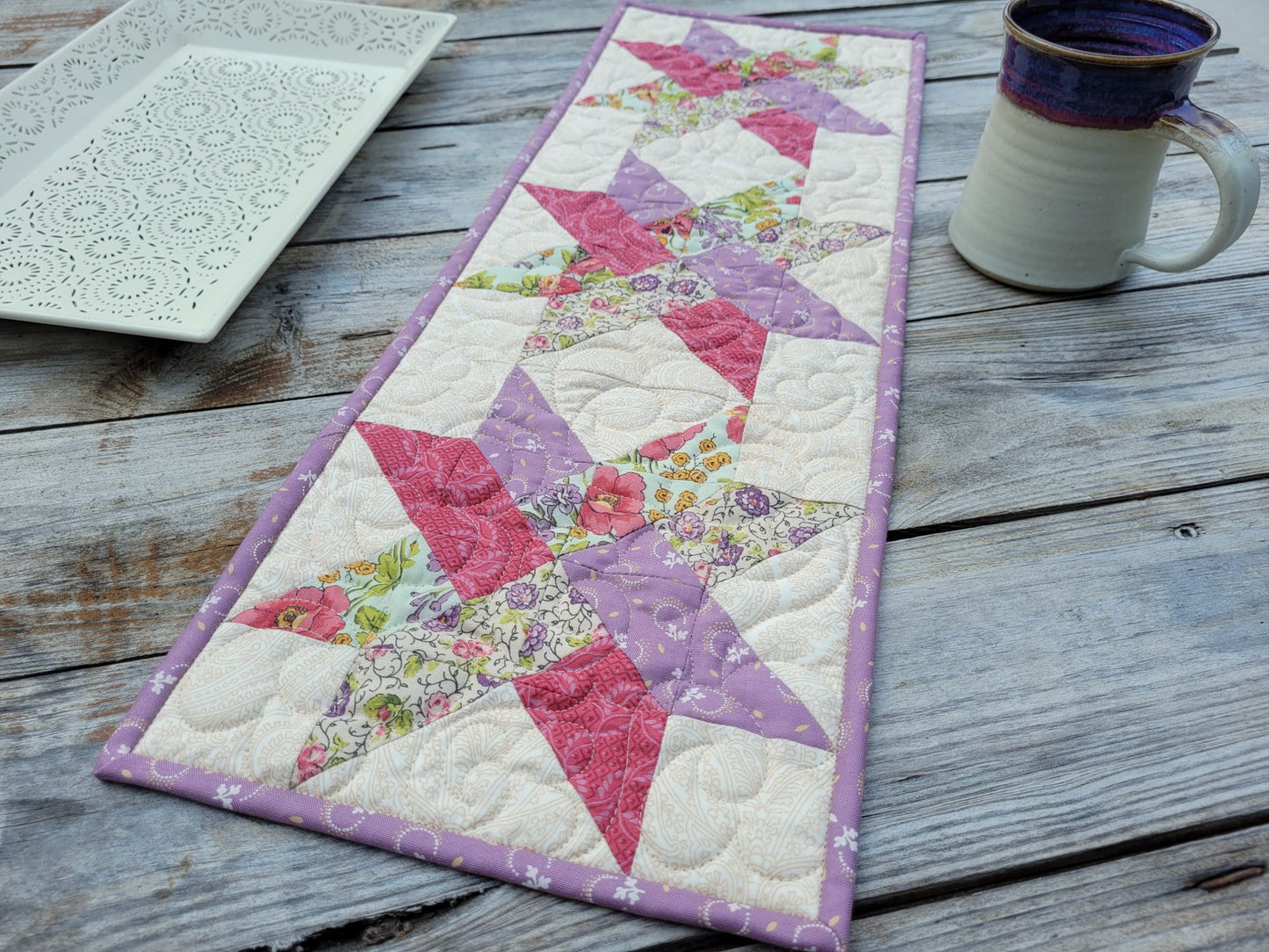 A handmade quilted table runner with a floral woven star pattern, styled on a rustic wooden table next to a ceramic mug and a white decorative tray.