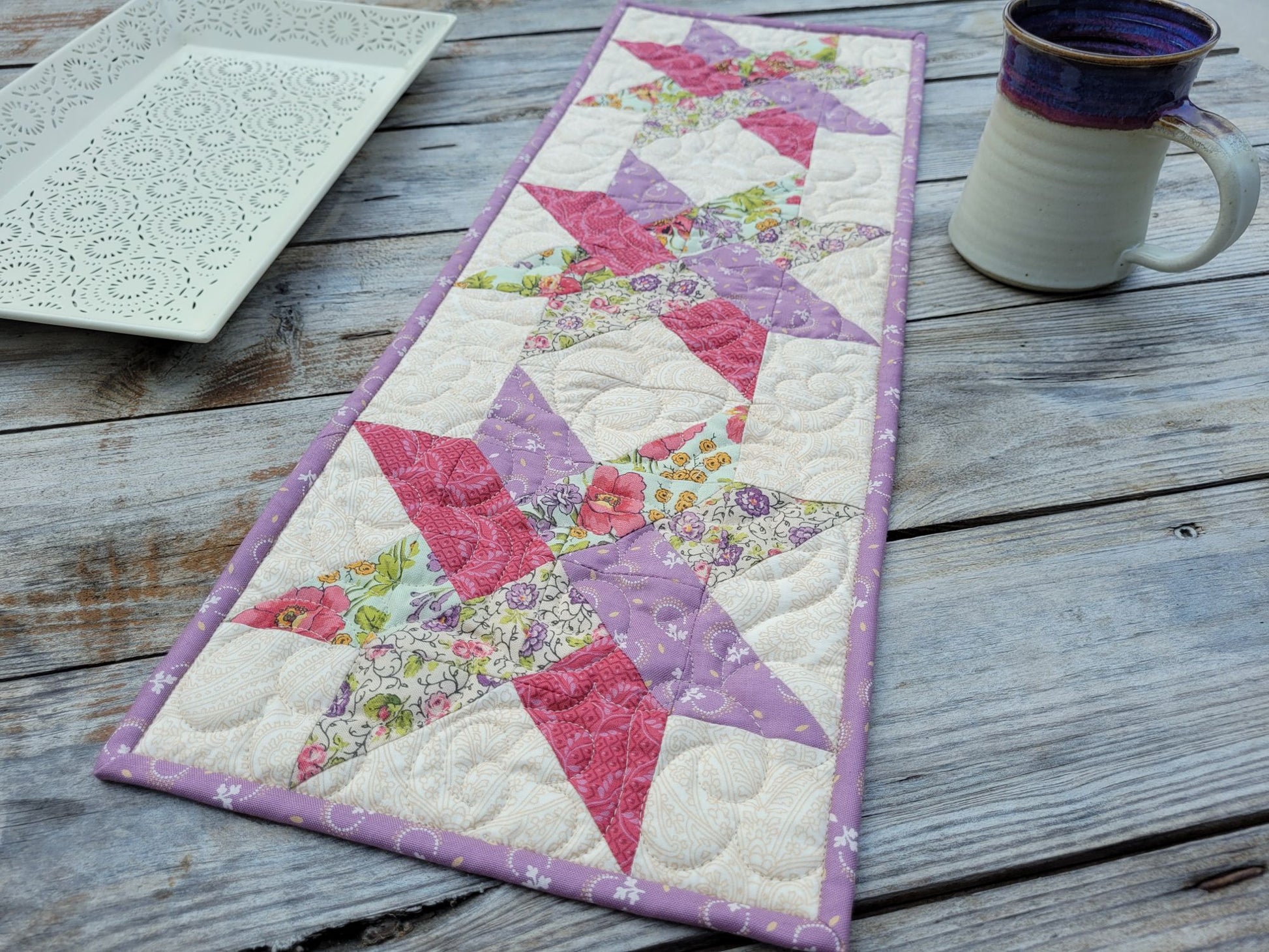 A handmade quilted table runner with a floral woven star pattern, styled on a rustic wooden table next to a ceramic mug and a white decorative tray.