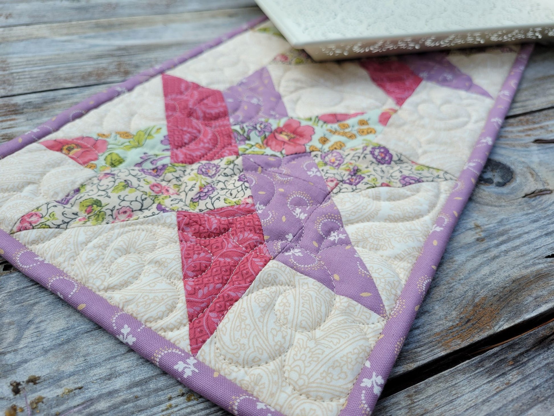 A close-up of a handmade quilted table runner featuring a woven star patchwork design in pink, purple, and floral prints, laid out on a rustic weathered wood surface.