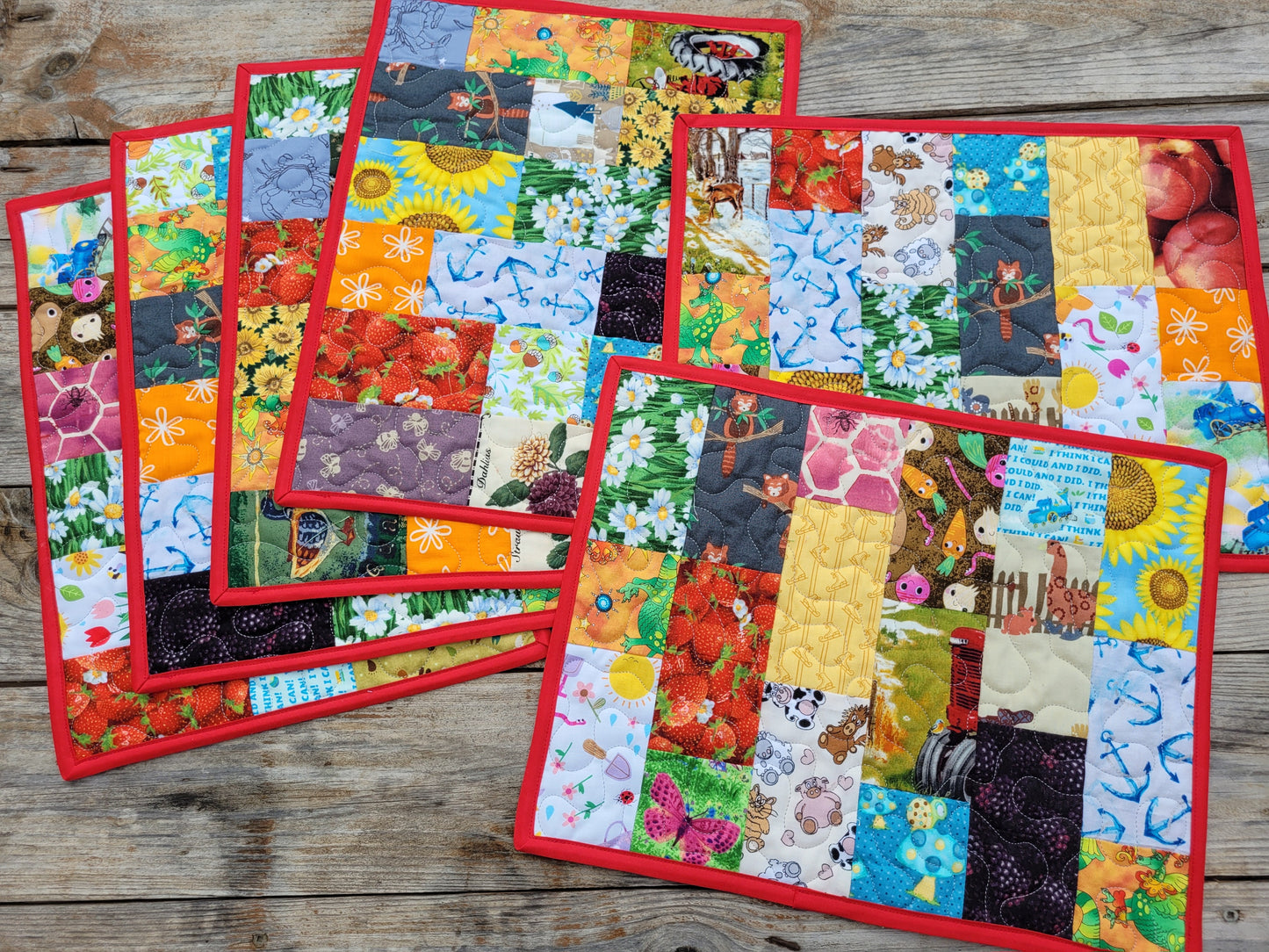 A set of colorful patchwork quilted placemats with red borders, arranged on a rustic wooden tabletop. Each placemat features a unique multicolor patterned design including floral, nature, and whimsical prints.