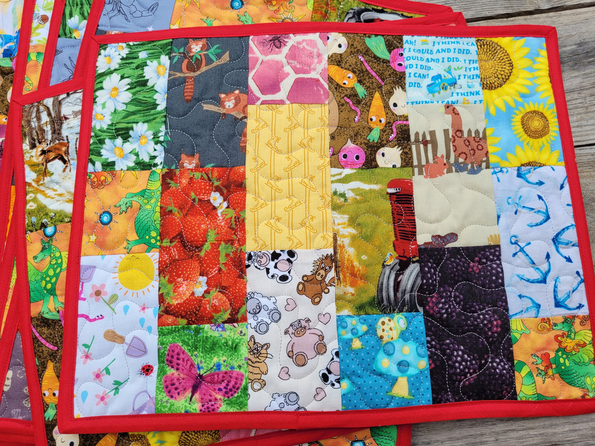 A close-up of a colorful quilted patchwork mat with a red border, featuring various animal, nature, and patterned fabric squares, shown on a wooden surface.
